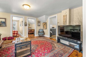 A living room with a large flat-screen TV, a wooden chest, a red patterned rug, and a dining area with a table and chairs.