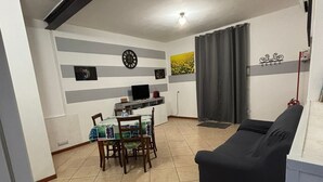 Un salon avec un canapé foncé, une table à manger avec des chaises, un téléviseur et une horloge au mur.