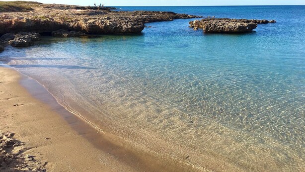 Uma praia com águas cristalinas e rasas, formações rochosas e um areal arenoso.