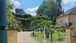Une entrée fermée par un portail, avec une allée gravillonnée menant à une maison dotée d’un mur en pierre et d’un portail en métal.