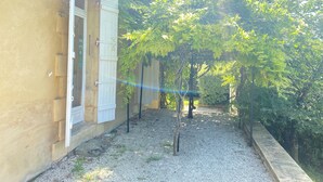 Une cour avec un chemin de gravier, une pergola couverte de plantes grimpantes et un bâtiment doté d’une porte et de volets.