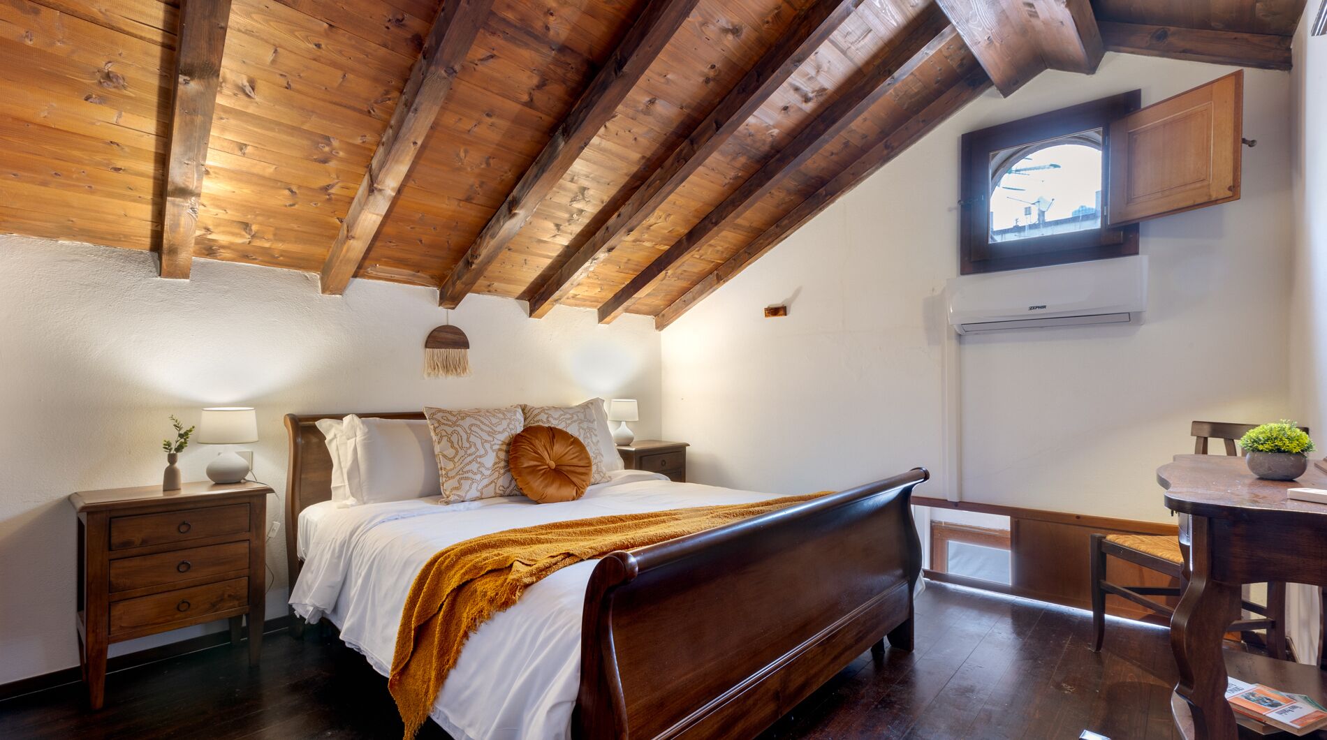 A bedroom with a wooden ceiling, a bed with white bedding, a bedside table, a chair, and a small table with a plant.