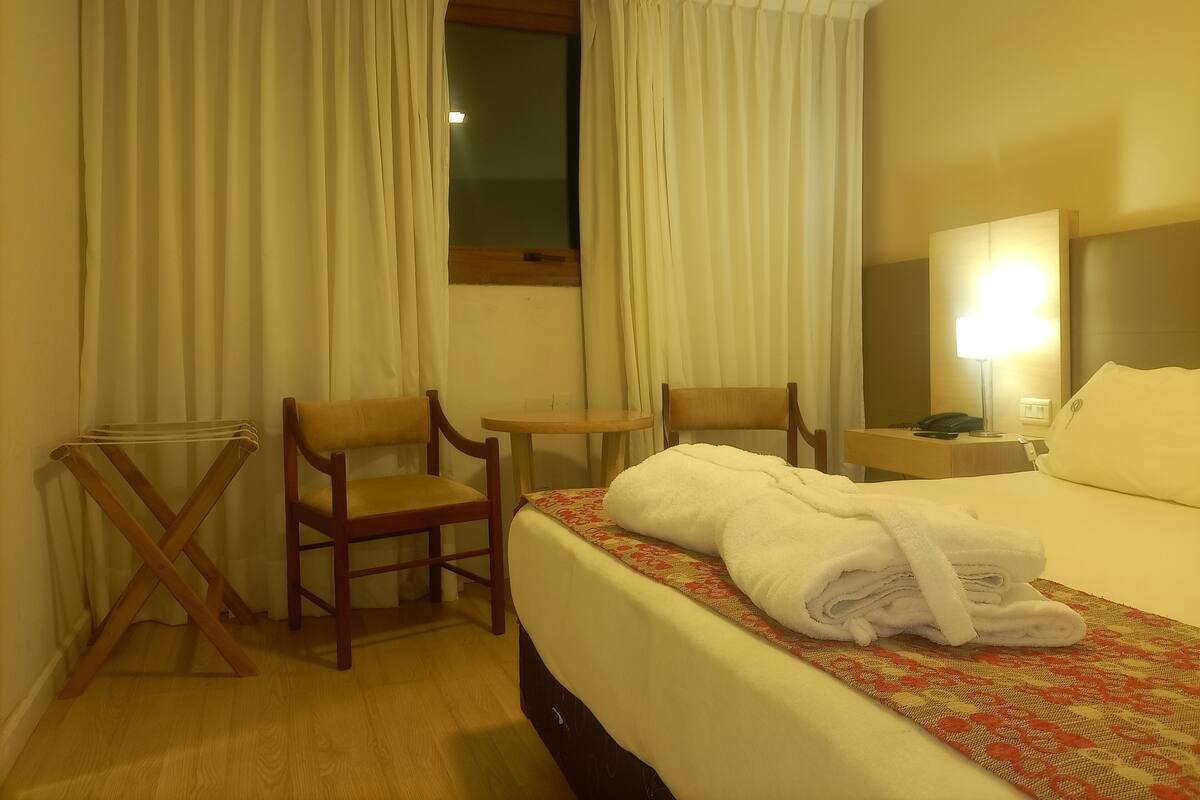 Ein Hotelzimmer mit einem Bett, zwei Stühlen, einem kleinen Tisch, einem Nachttisch mit einer Lampe und Vorhängen.