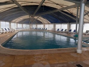 An indoor swimming pool with a tiled floor, surrounded by lounge chairs and a covered roof structure.