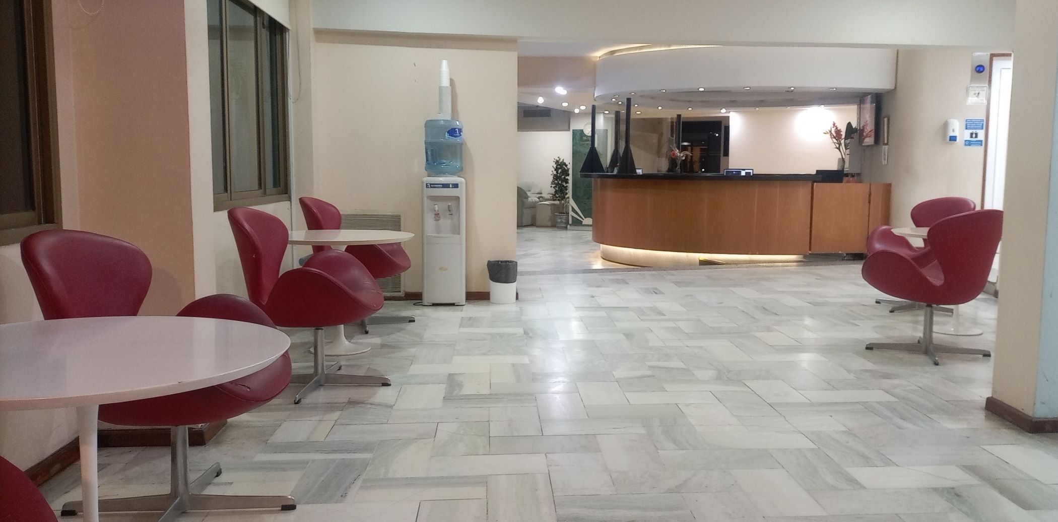 A hotel lobby with red chairs and tables, a reception desk, and a water dispenser.