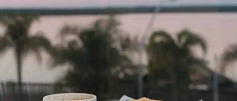 Mesa com xícara e prato de comida, com vista para um corpo d'água e palmeiras ao fundo.