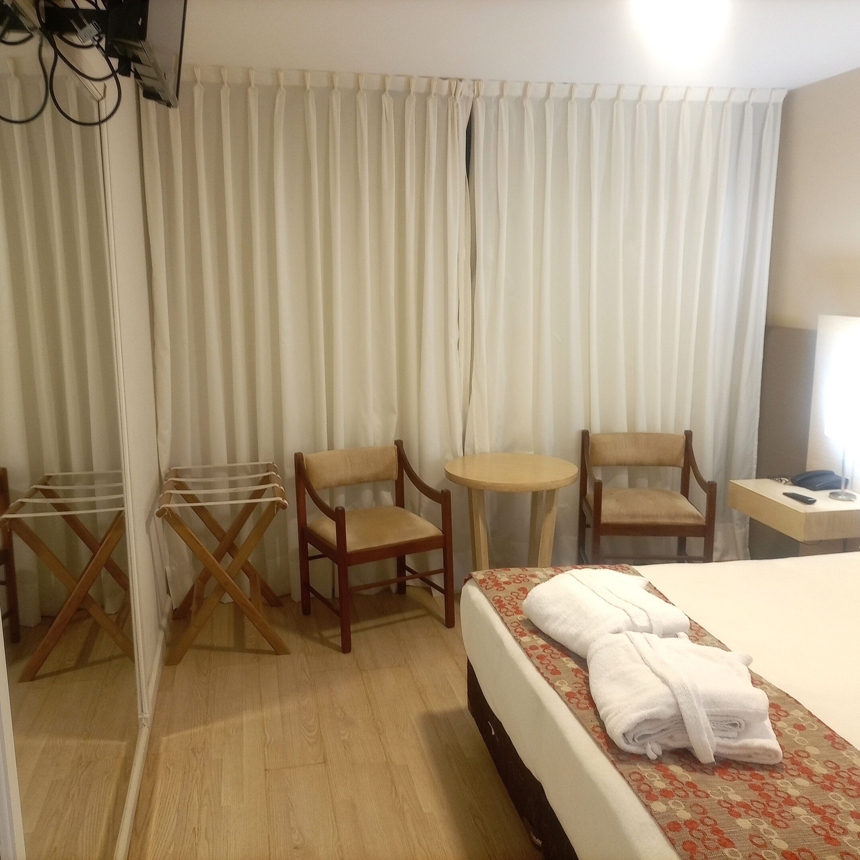 Habitación de hotel con cama, dos sillas de madera, una mesita redonda, un tendedero y un televisor montado en la pared.