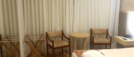 Habitación de hotel con cama, dos sillas de madera, una mesita redonda, un tendedero y un televisor montado en la pared.