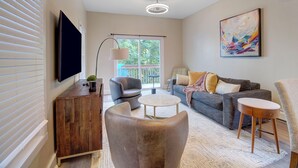 Een moderne woonkamer met een flatscreen televisie, een houten kast, een grijze bank met kussens, een ronde salontafel en een schilderij aan de muur.