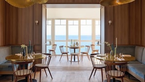 A dining area with round tables and chairs, wooden walls, and a view of the sea. - Les Bassans - Fontenille Collection (Perros-Guirec)