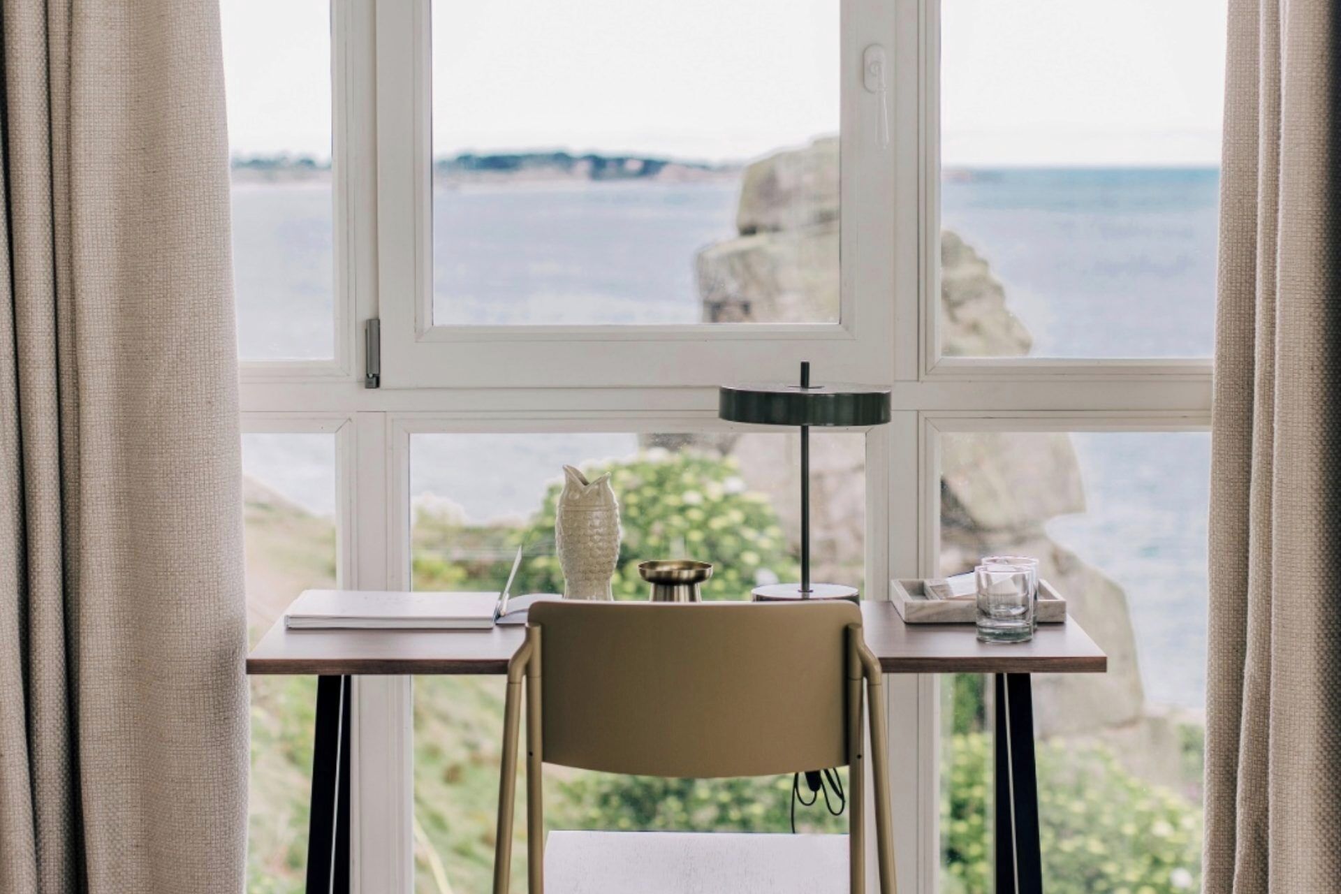 A room with a view of the sea through large windows, a desk with a chair, and a lamp.