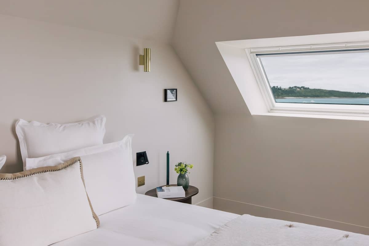 A bedroom with a bed, a nightstand, a vase with flowers, a picture on the wall, and a window with a view of a body of water.