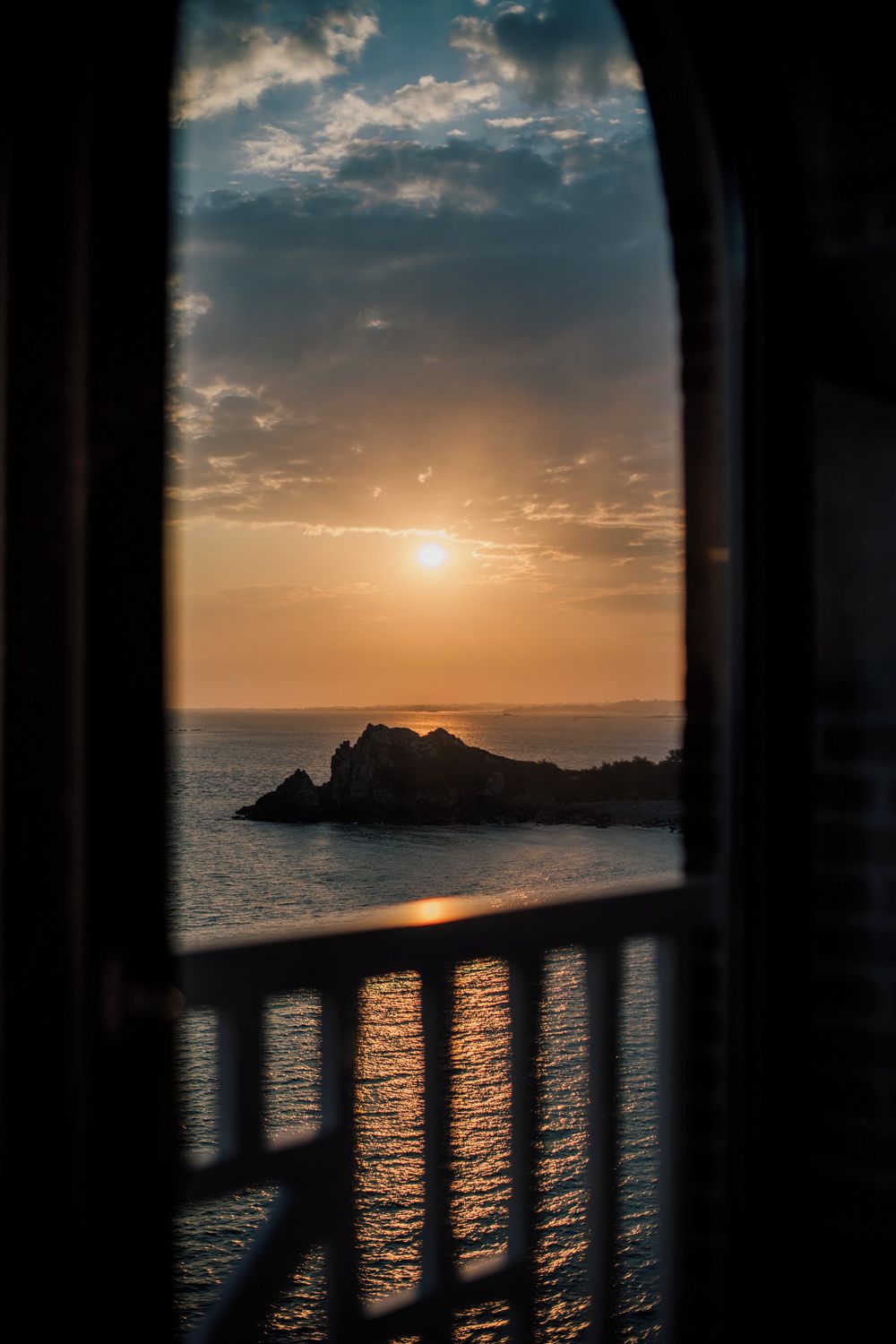 A sunset view through a window with a railing, overlooking a body of water and a distant landmass.