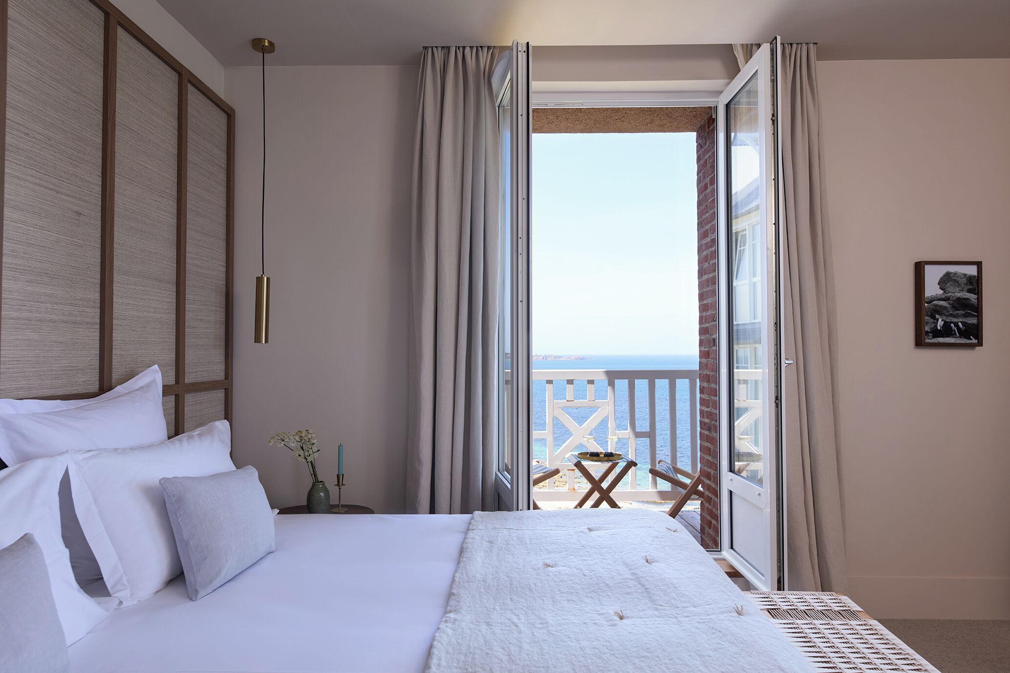 A bedroom with a bed, a nightstand, a vase with flowers, a chair, and a view of the sea through an open door.