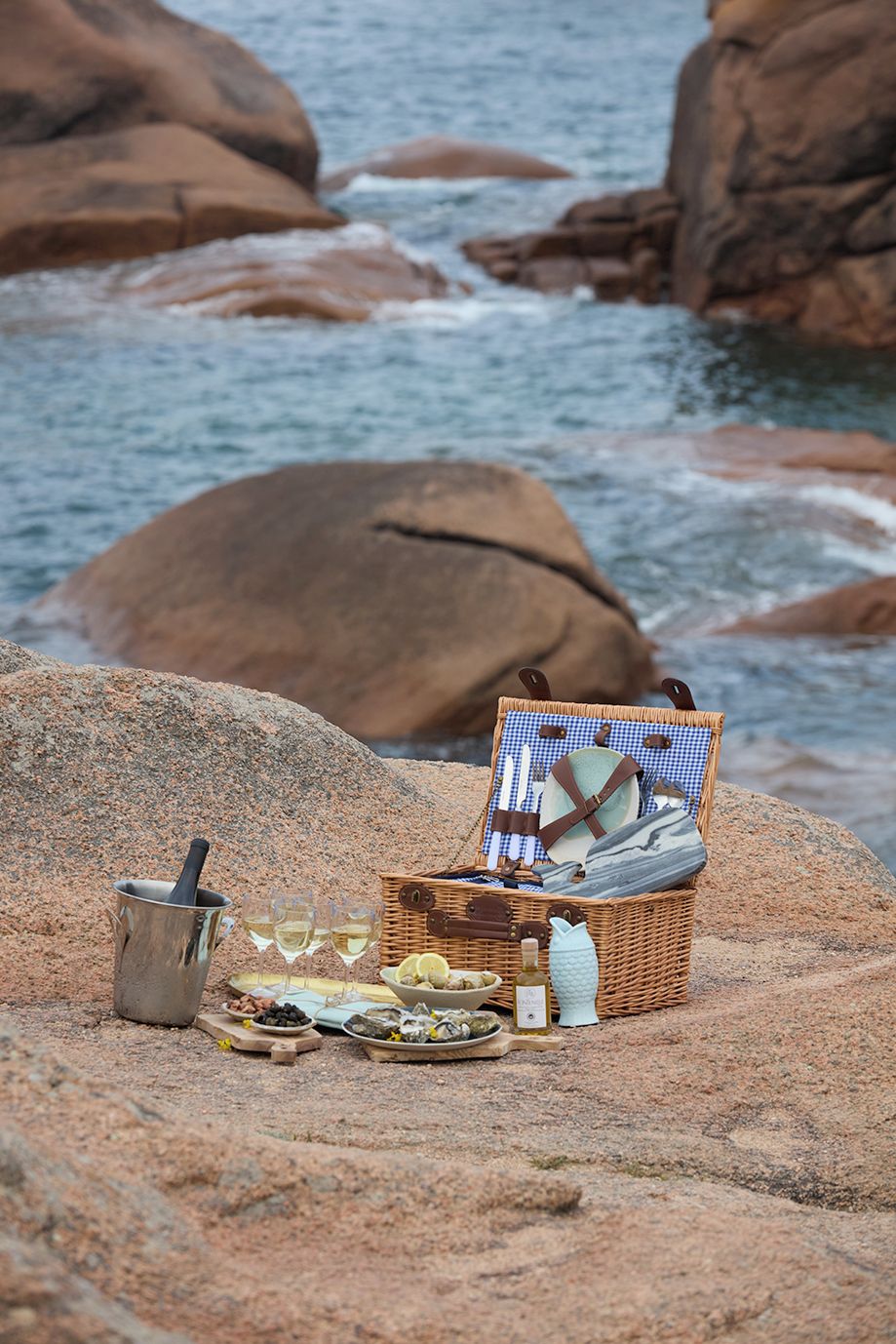 A picnic setup with a basket, wine bottle, glasses, and food on a rocky shore.