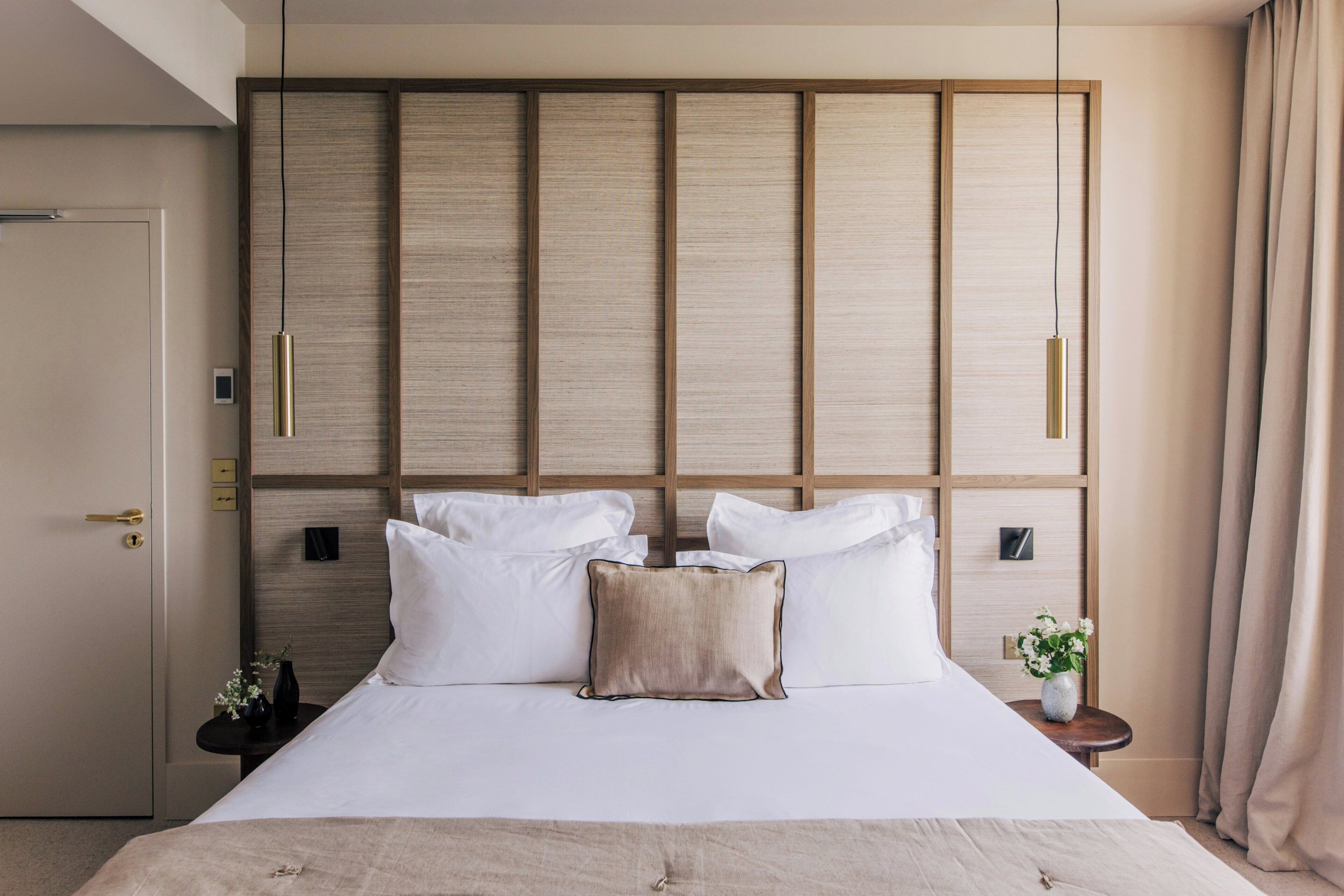 Una habitación de hotel con una cama grande, un cabecero de madera, una mesita de noche con un florero y una puerta con picaporte.