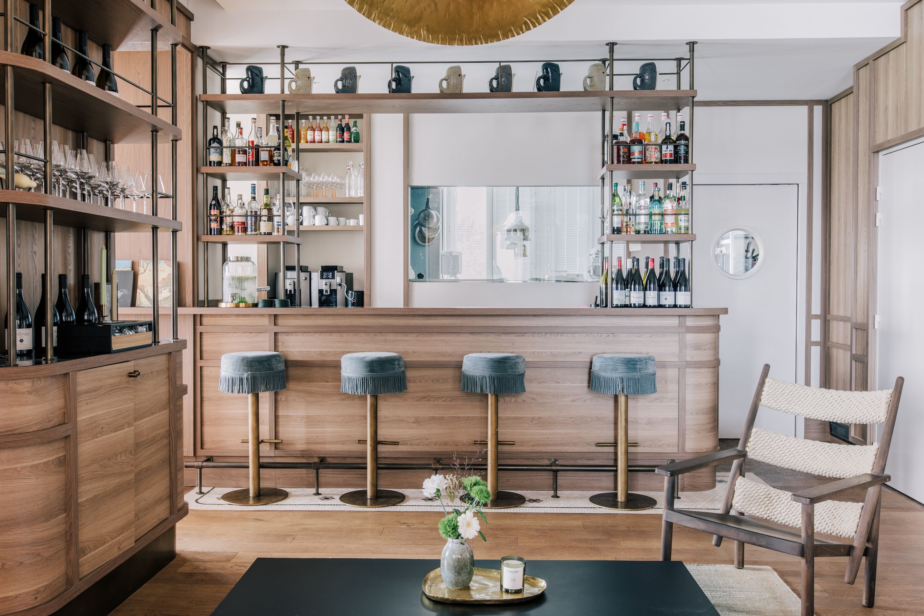 A bar area with multiple shelves stocked with bottles, glasses, and a coffee machine. There are four stools with blue cushions and a chair with a woven seat. A table with a vase and candle is in the foreground.