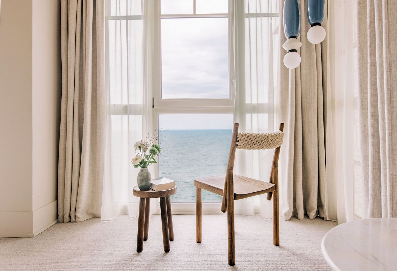 A room with a view of the sea, a wooden chair, a small table with a vase, and a round table.