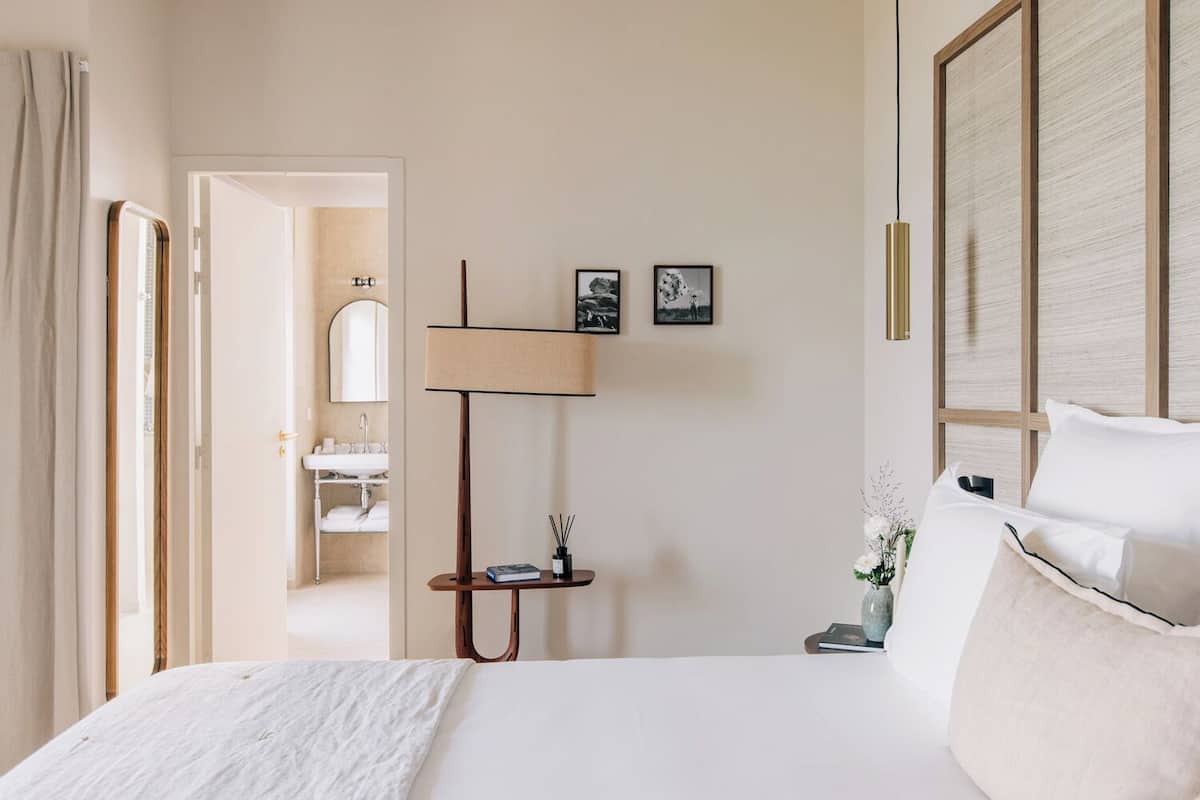 A hotel room with a bed, a bedside table with a lamp, a small table with a vase, and a mirror.