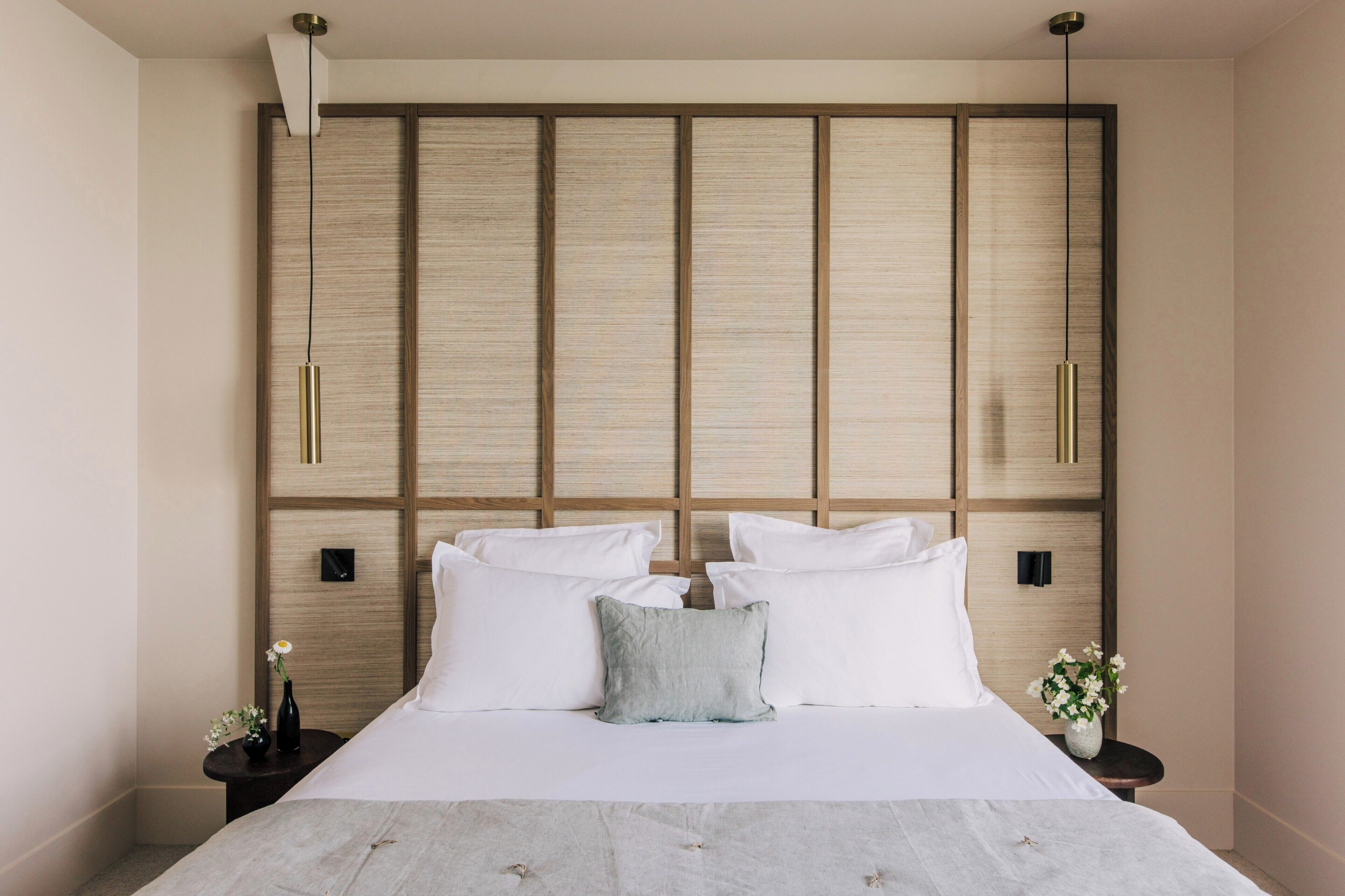 A modern hotel room with a large bed, a wooden headboard, two bedside tables, and a small nightstand with a vase of flowers.