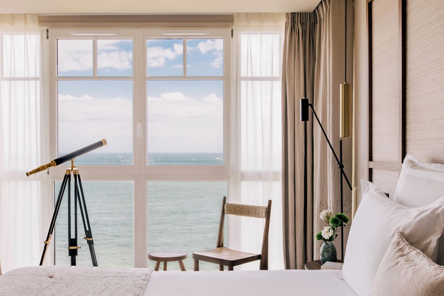 Una habitación de hotel con un ventanal grande, un telescopio, una silla, una mesita y una cama.