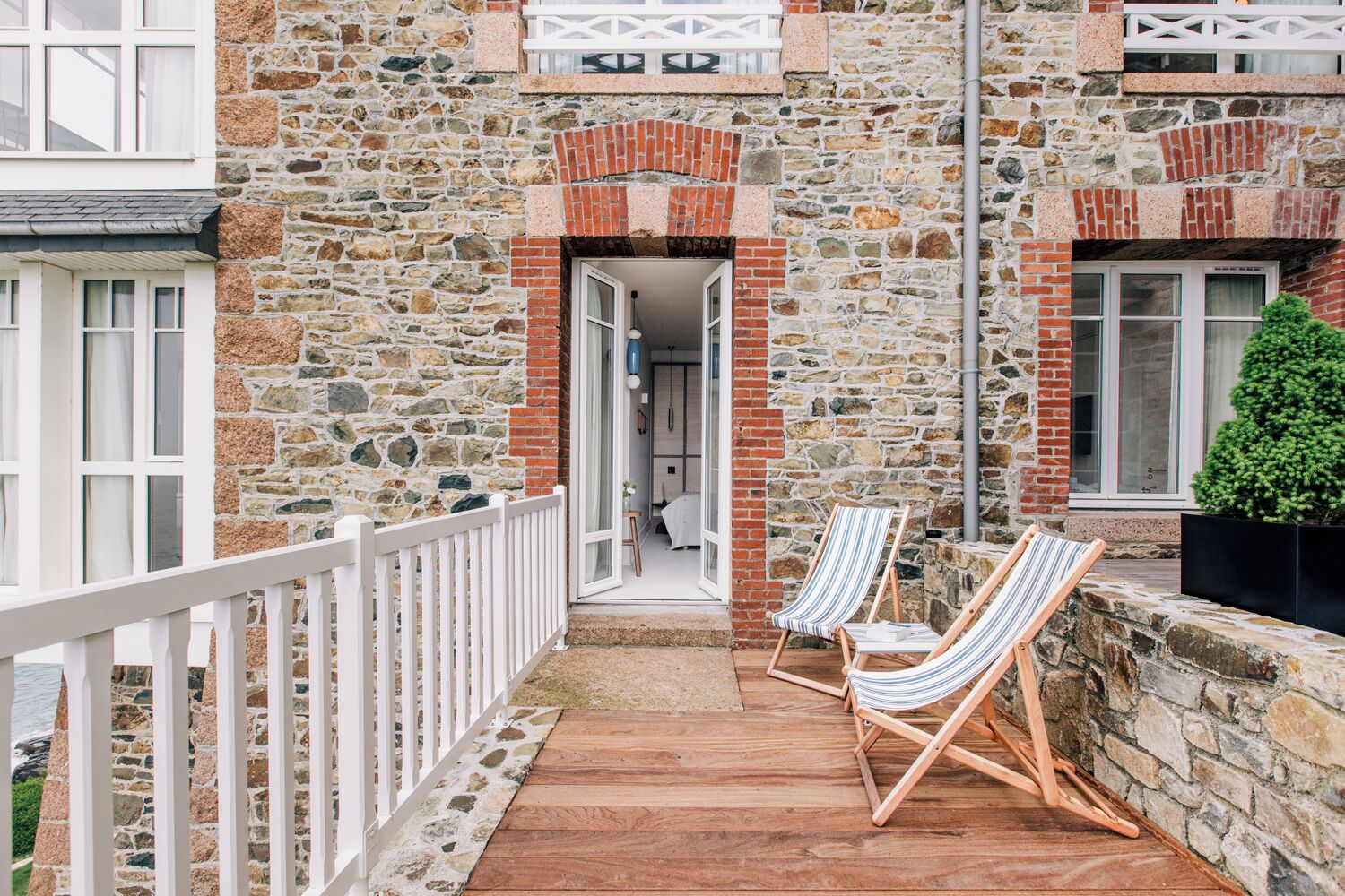 Un bâtiment en pierre doté d’une terrasse en bois, de deux chaises longues à rayures et d’une rambarde blanche.
