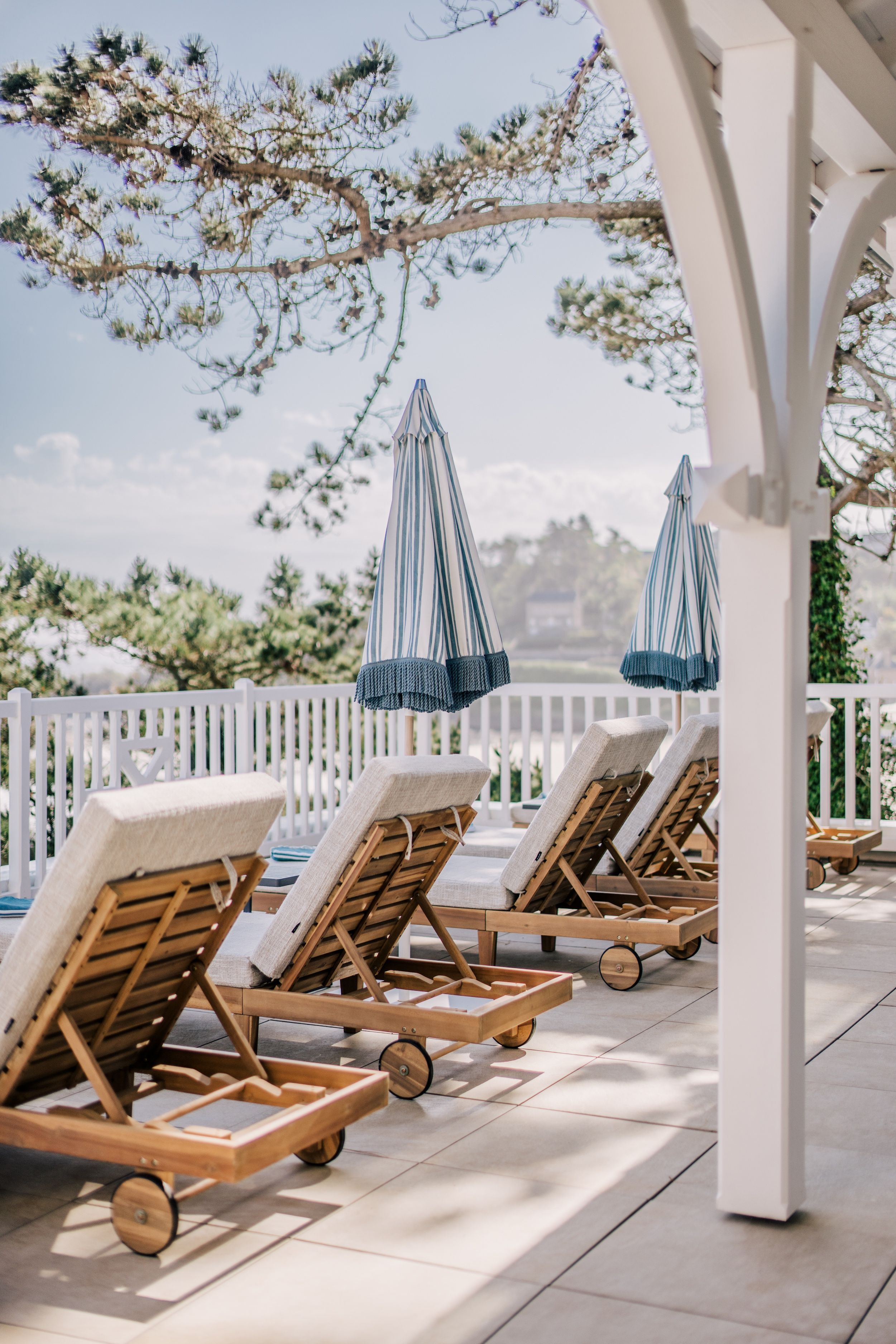 Eine Reihe von Holz-Loungestühlen mit weißen Kissen, angeordnet auf einer gefliesten Terrasse unter einer weißen Pergola mit blauen Streifen an den Sonnenschirmen.