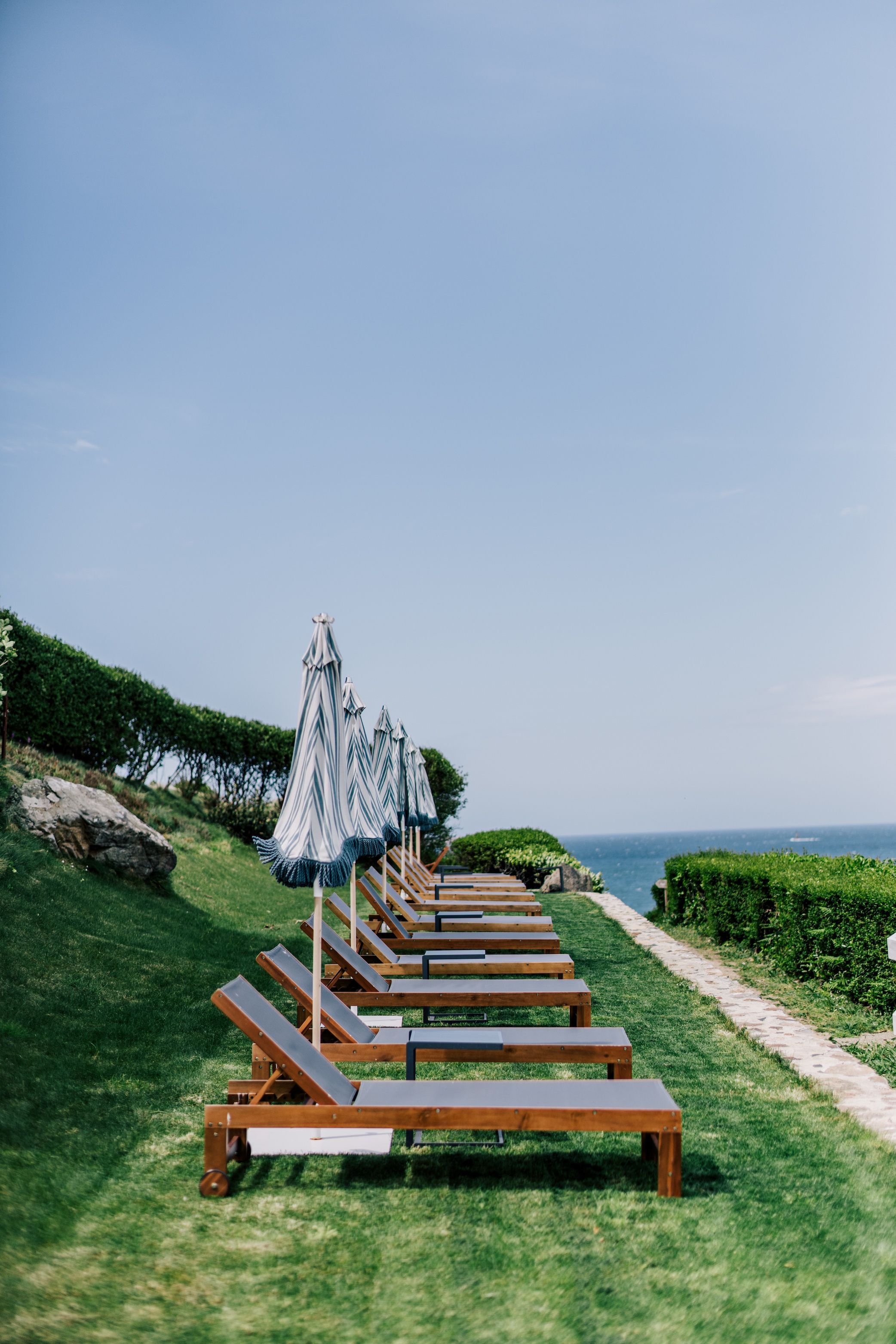 A row of wooden lounge chairs with striped umbrellas, arranged on a grassy area with a clear view of the sea.