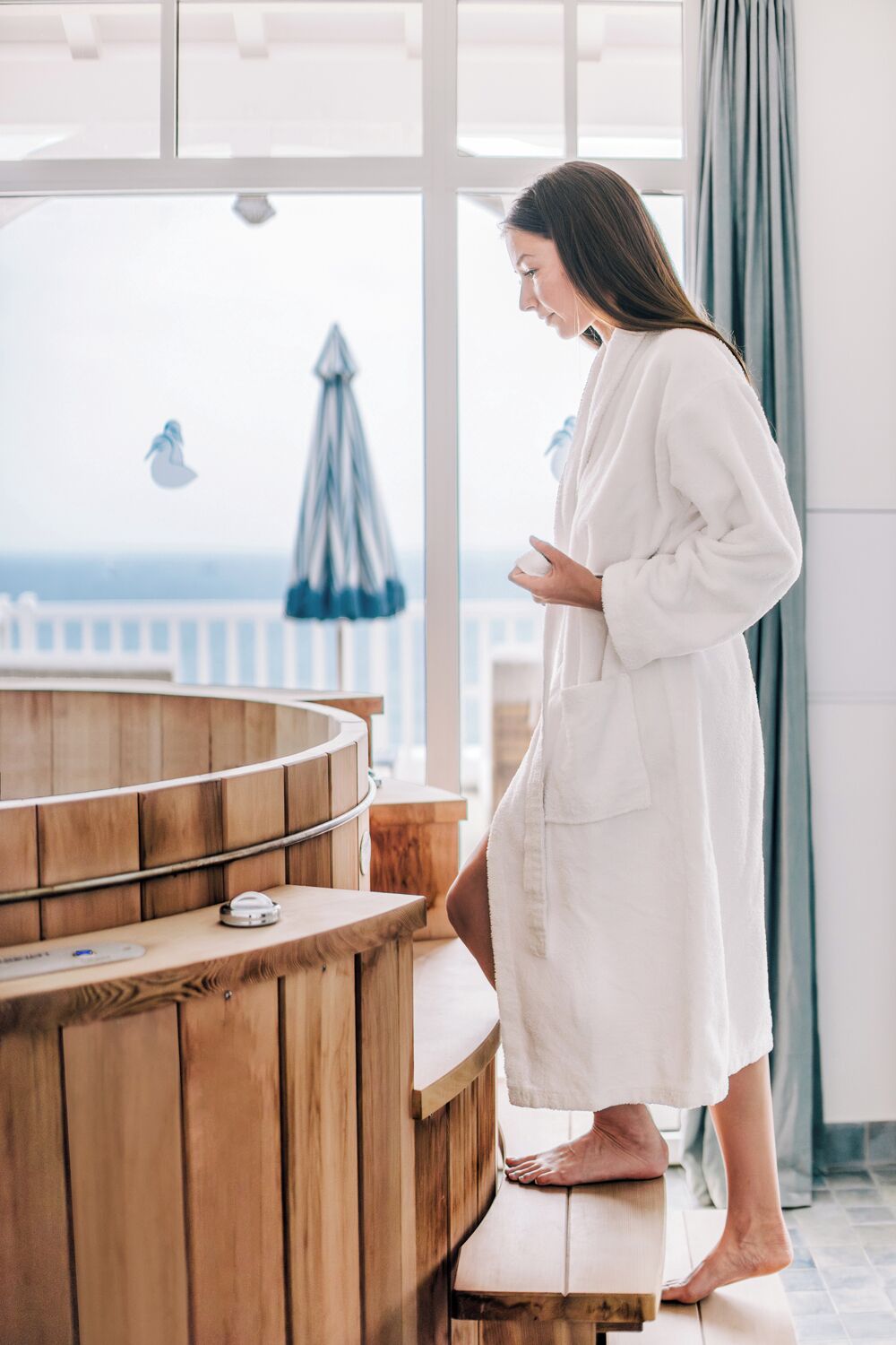 A person in a white bathrobe standing by a wooden sauna with a view of the sea.