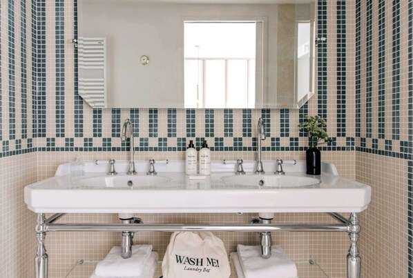 A bathroom with a large white sink, multiple faucets, and a mirror above. The walls are tiled with a pattern of small squares in shades of green and beige. There is a towel rack with two towels and a bag labeled "WASH ME!" on the floor. - Les Bassans - Fontenille Collection (Perros-Guirec)