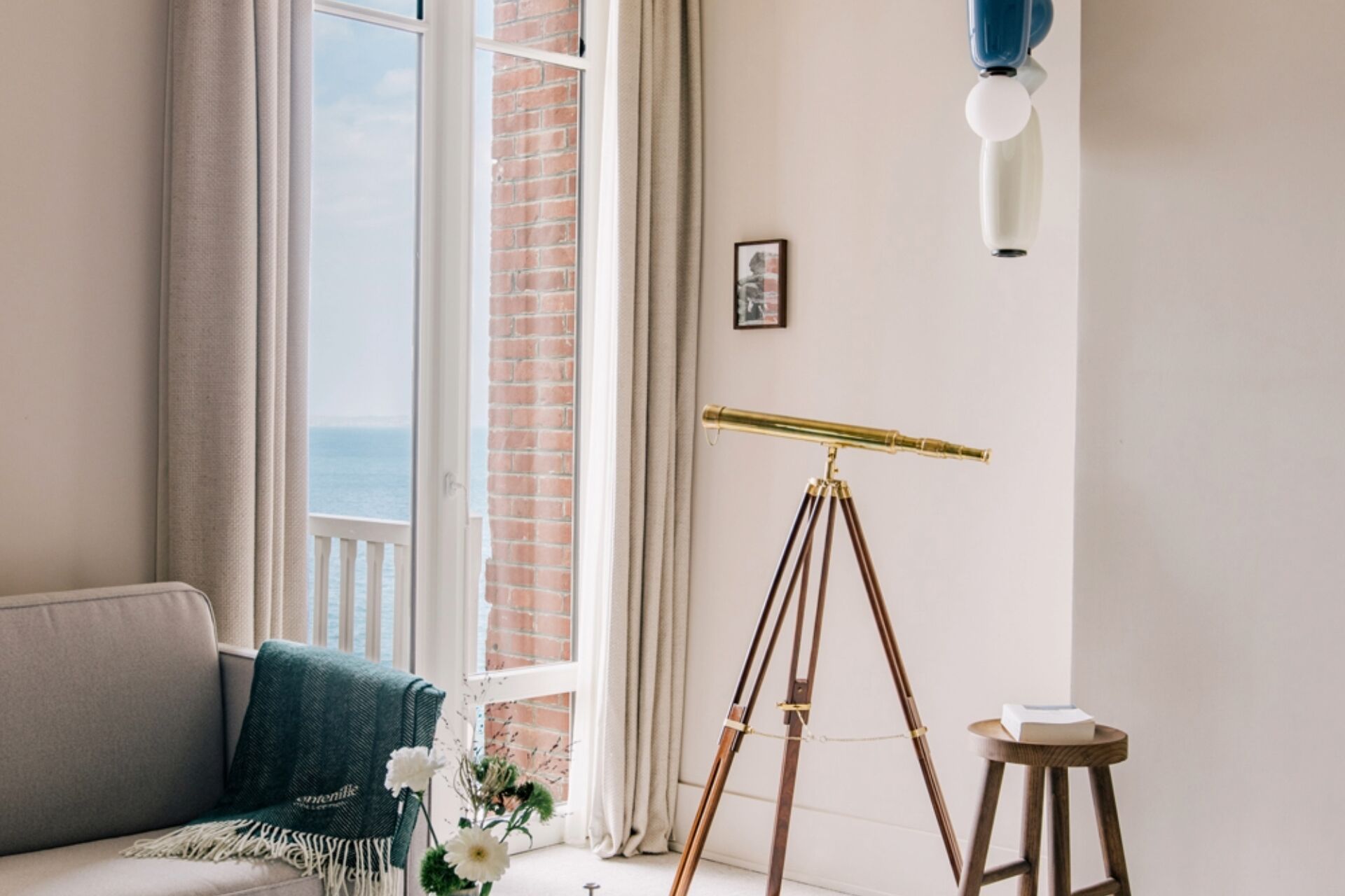 Una habitación con un telescopio sobre un trípode, un sofá con una manta, un taburete pequeño con un libro y un cuadro en la pared.