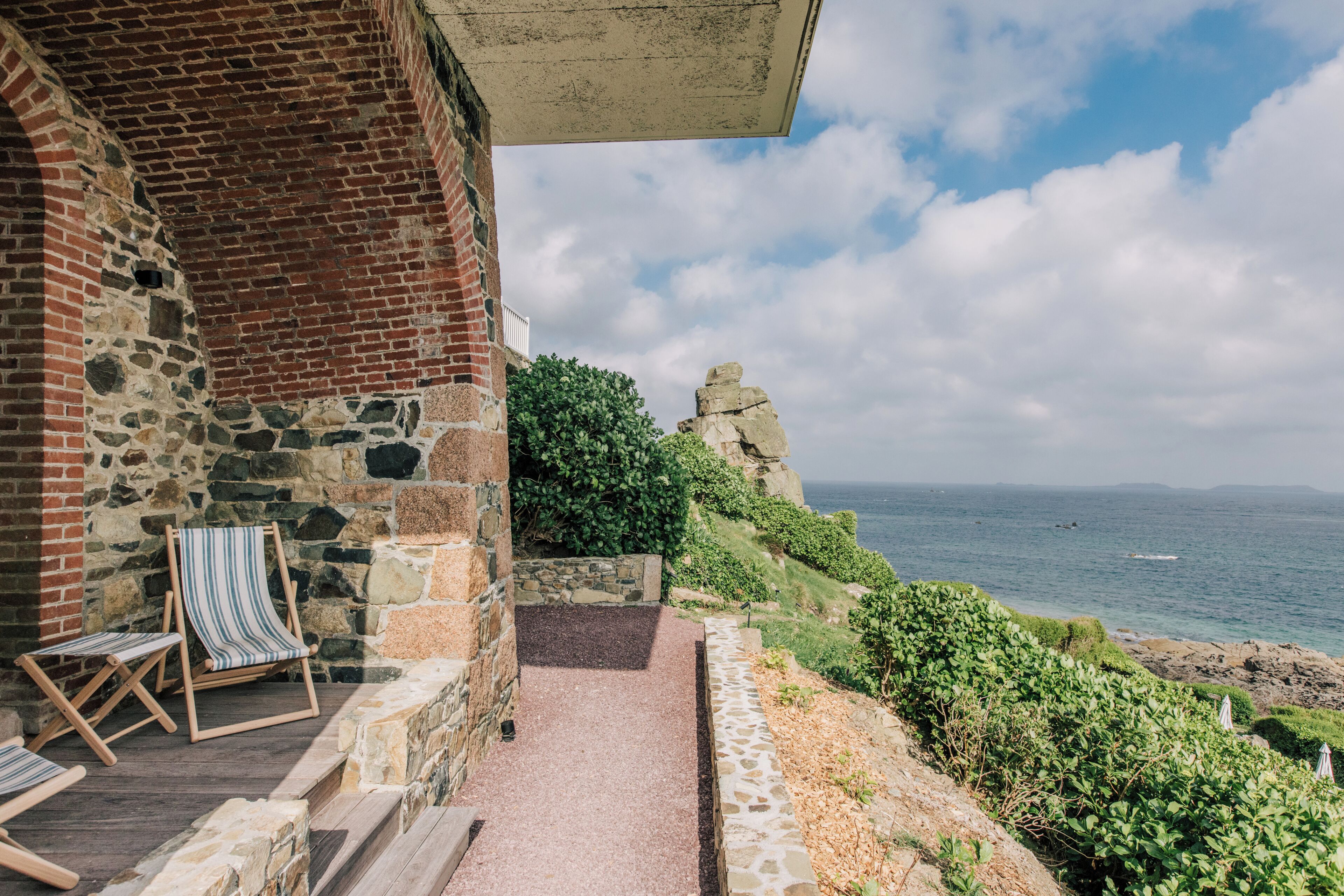 A stone building with a brick section, a pathway leading to a seating area with striped chairs, and a view of the sea.
