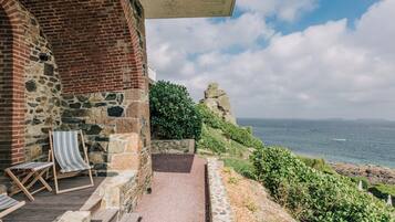 A stone building with a brick section, a pathway leading to a seating area with striped chairs, and a view of the sea.
