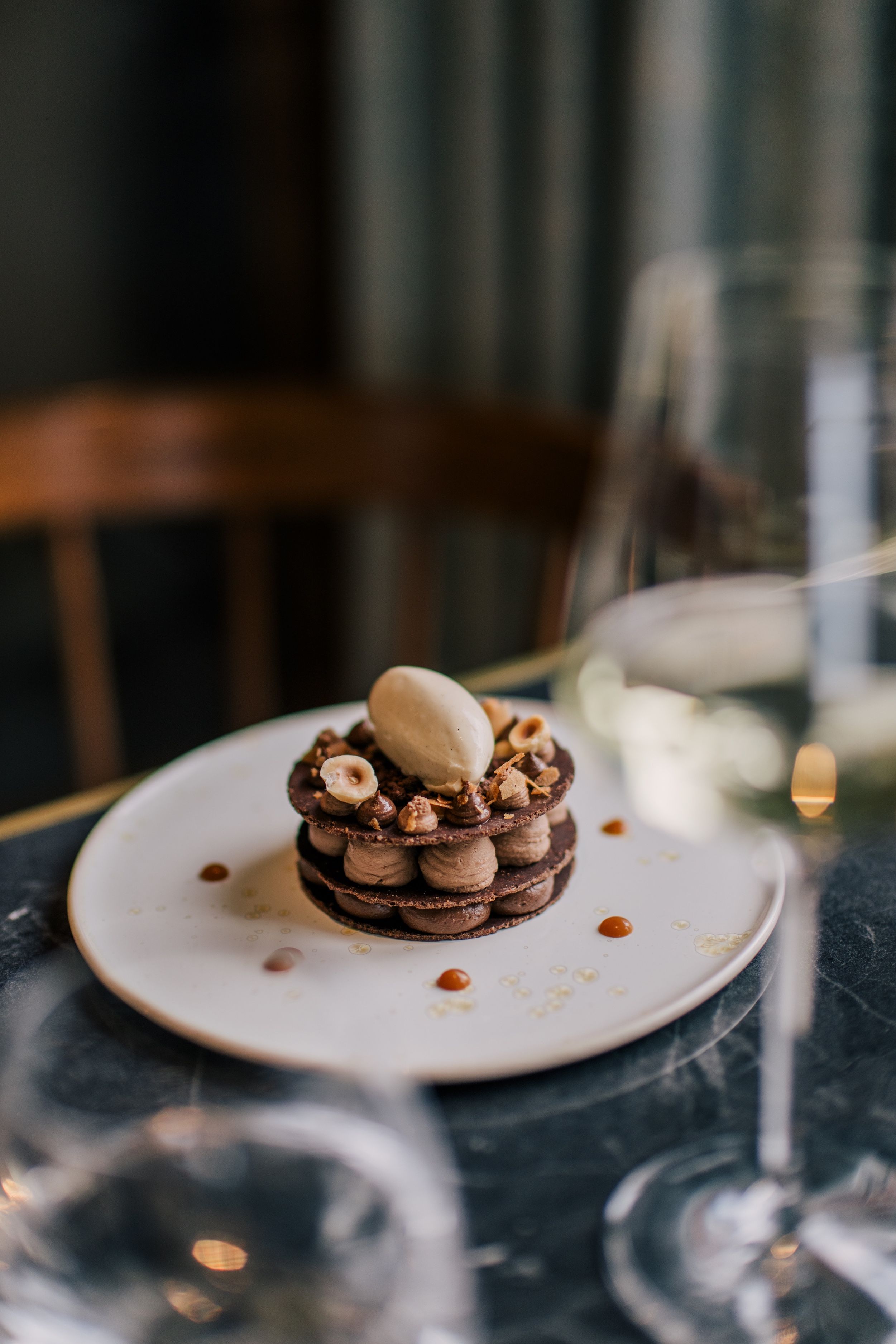 A dessert on a plate with a chocolate base, hazelnuts, and a scoop of ice cream.