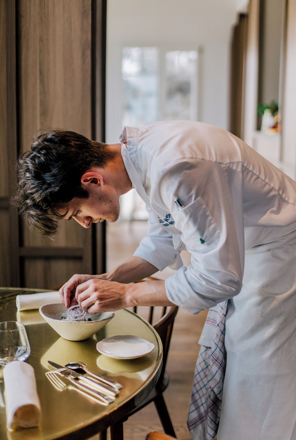 A person in a white chef's uniform preparing food at a table with a bowl, plate, and cutlery.