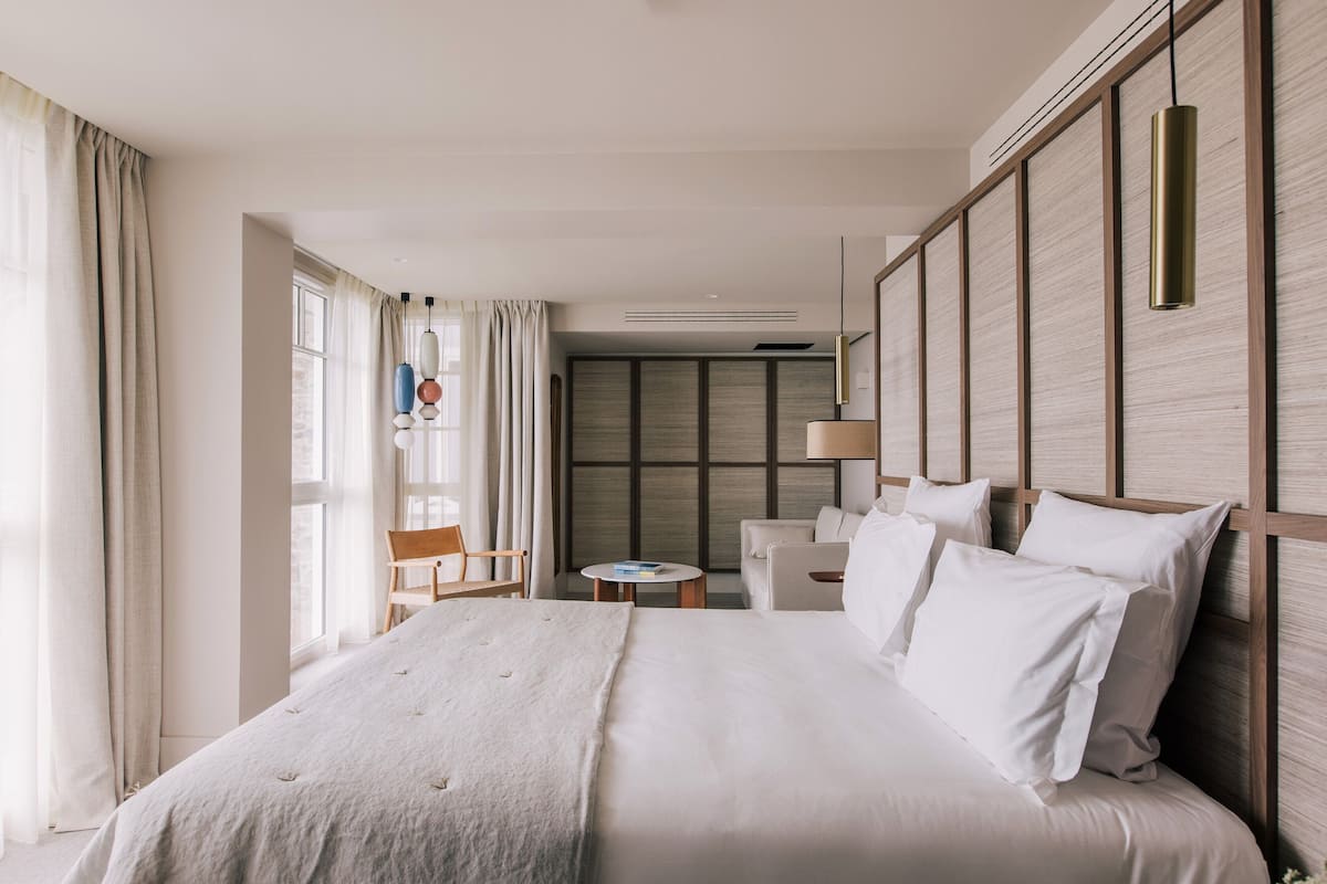 A modern hotel room with a large bed, a small table, a chair, and a view of the city through large windows.