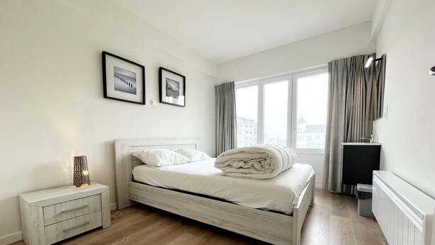Quarto com cama, mesa de cabeceira e janela com cortinas.