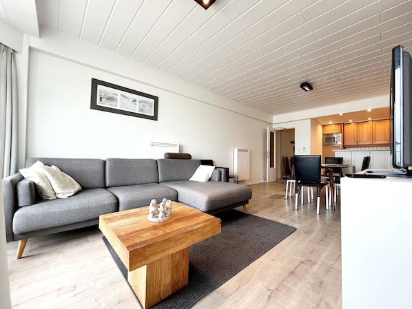 A modern living room with a grey sectional sofa, a wooden coffee table, and a dining area with a white table and chairs. - Apartment in Wenduine With Sea Views (De Haan)