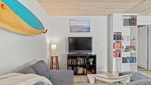 Una sala de estar con un sofá gris, una estantería, una tabla de surf y un televisor.