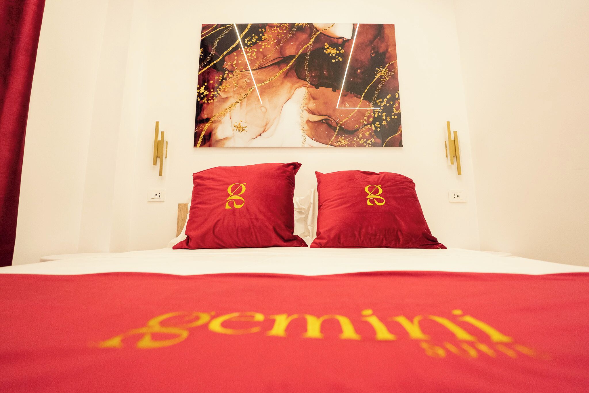 A hotel room with a bed, red bedding, a large painting on the wall, and two wall-mounted light fixtures.