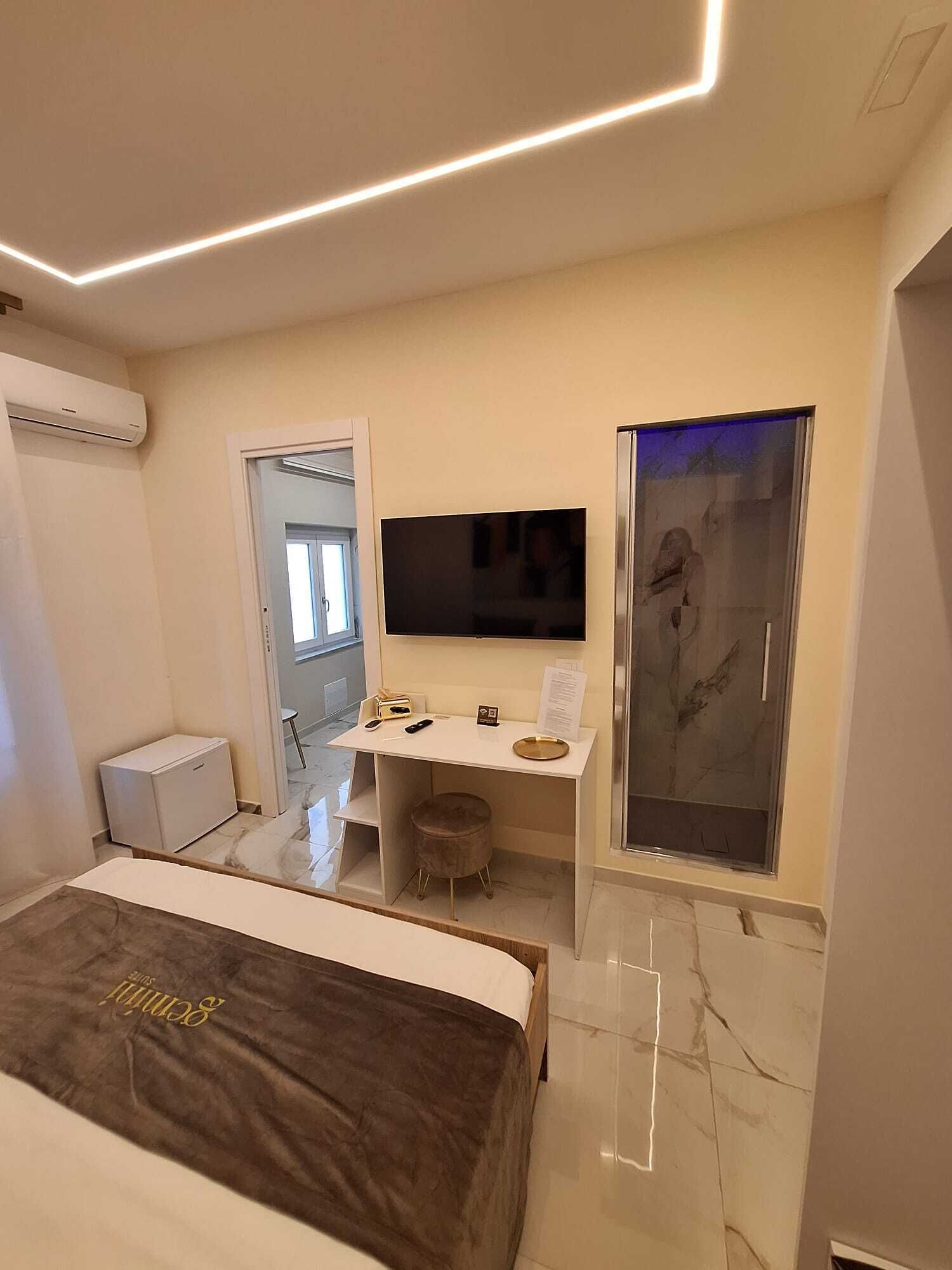 A modern hotel room with a bed, a desk with a flat-screen TV, a small stool, and a glass shower enclosure.