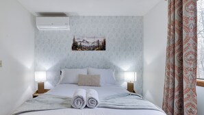 Una habitación de hotel con una cama bien hecha, dos lámparas de noche, un aire acondicionado montado en la pared y una ventana con cortinas estampadas.