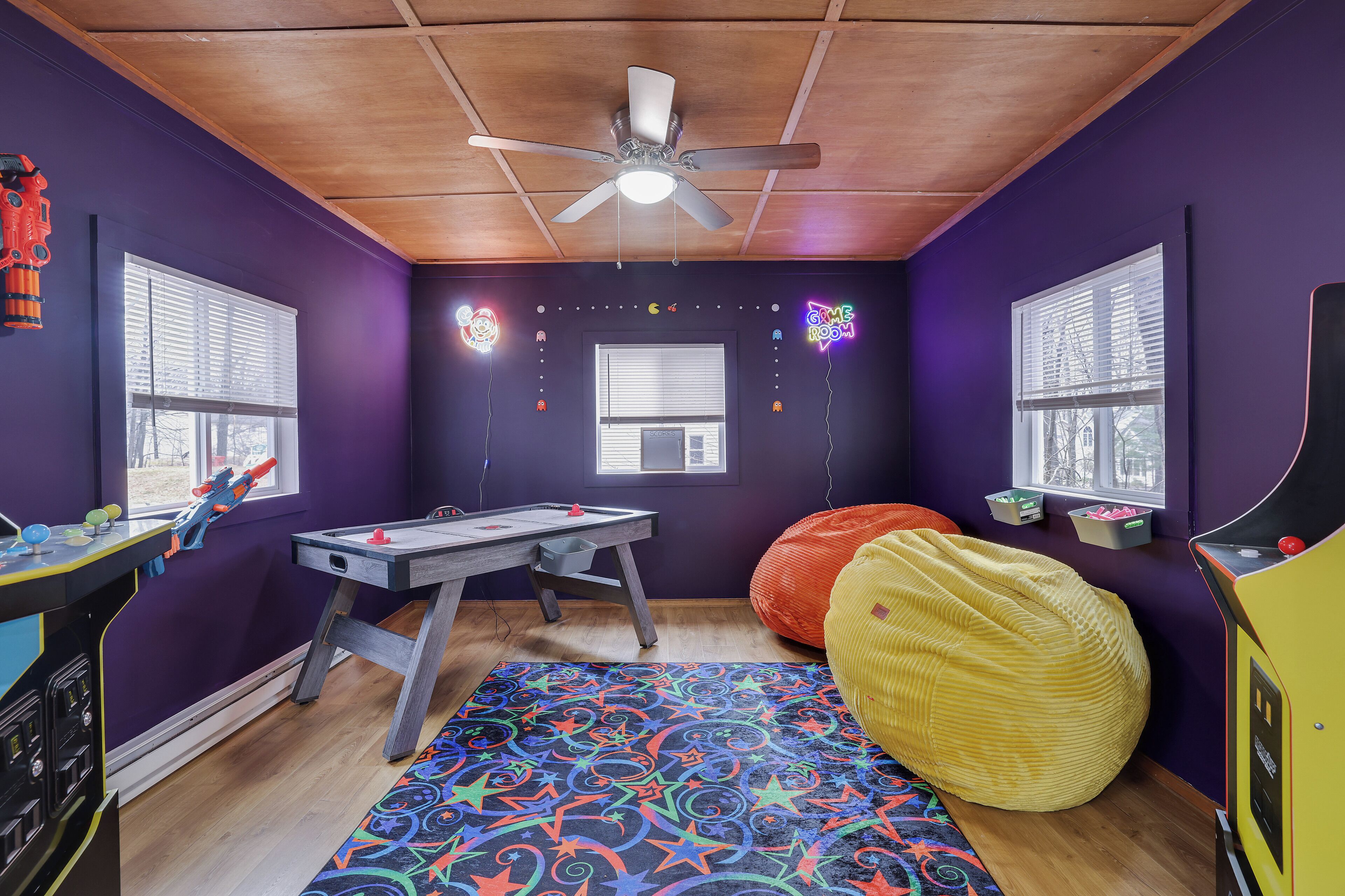 Une pièce aux murs violets, avec un plafond en bois, un ventilateur de plafond, un tapis coloré, un fauteuil en forme de poire, une table avec des jouets et des fenêtres équipées de stores.
