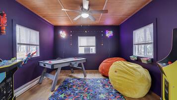 Une pièce aux murs violets, avec un plafond en bois, un ventilateur de plafond, un tapis coloré, un fauteuil en forme de poire, une table avec des jouets et des fenêtres équipées de stores.