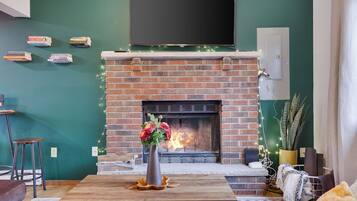 A cozy living room with a brick fireplace, a flat-screen TV mounted above it, and a table with a vase of flowers.