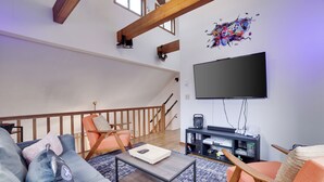 Una sala de estar con un sofá, una mesa de centro y dos sillones. Un televisor de pantalla plana está montado en la pared, y hay una escalera con vigas de madera.