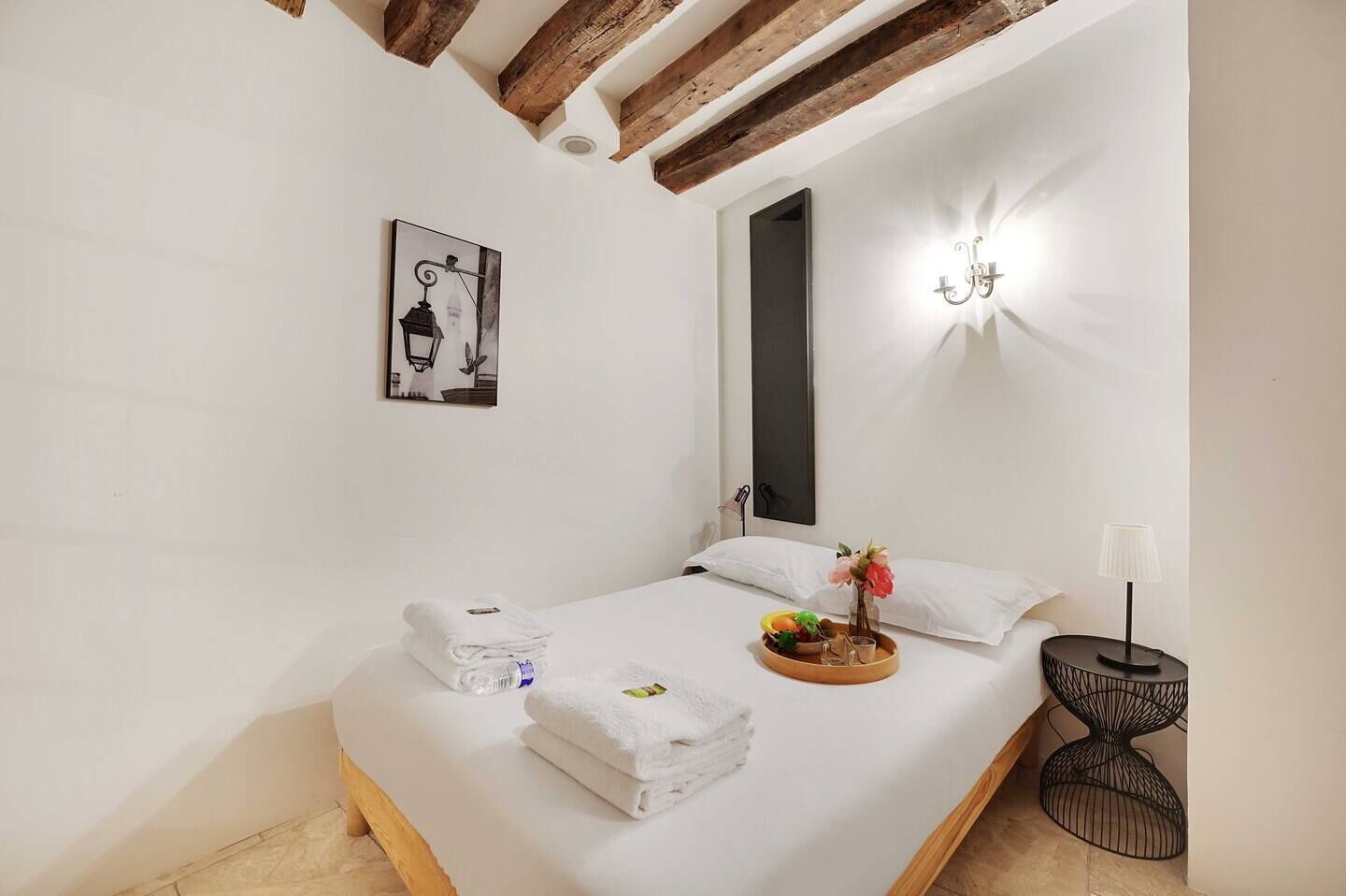 Una cama bien hecha con toallas, una bandeja de frutas y un jarrón de flores. Se observa una lámpara de pared y un cuadro en la pared.