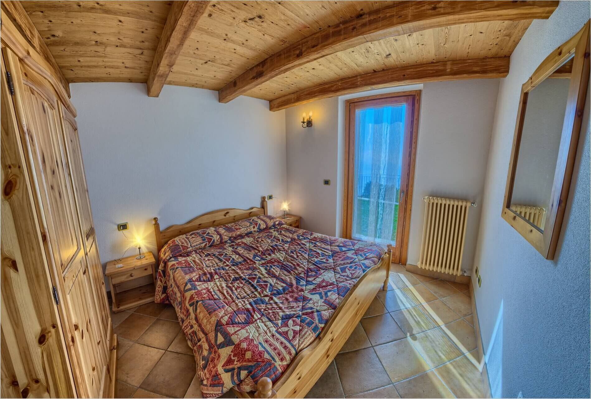 Una camera da letto con soffitto in legno, un letto con copriletto decorato, un comodino con lampada, una finestra con tende e un termosifone.