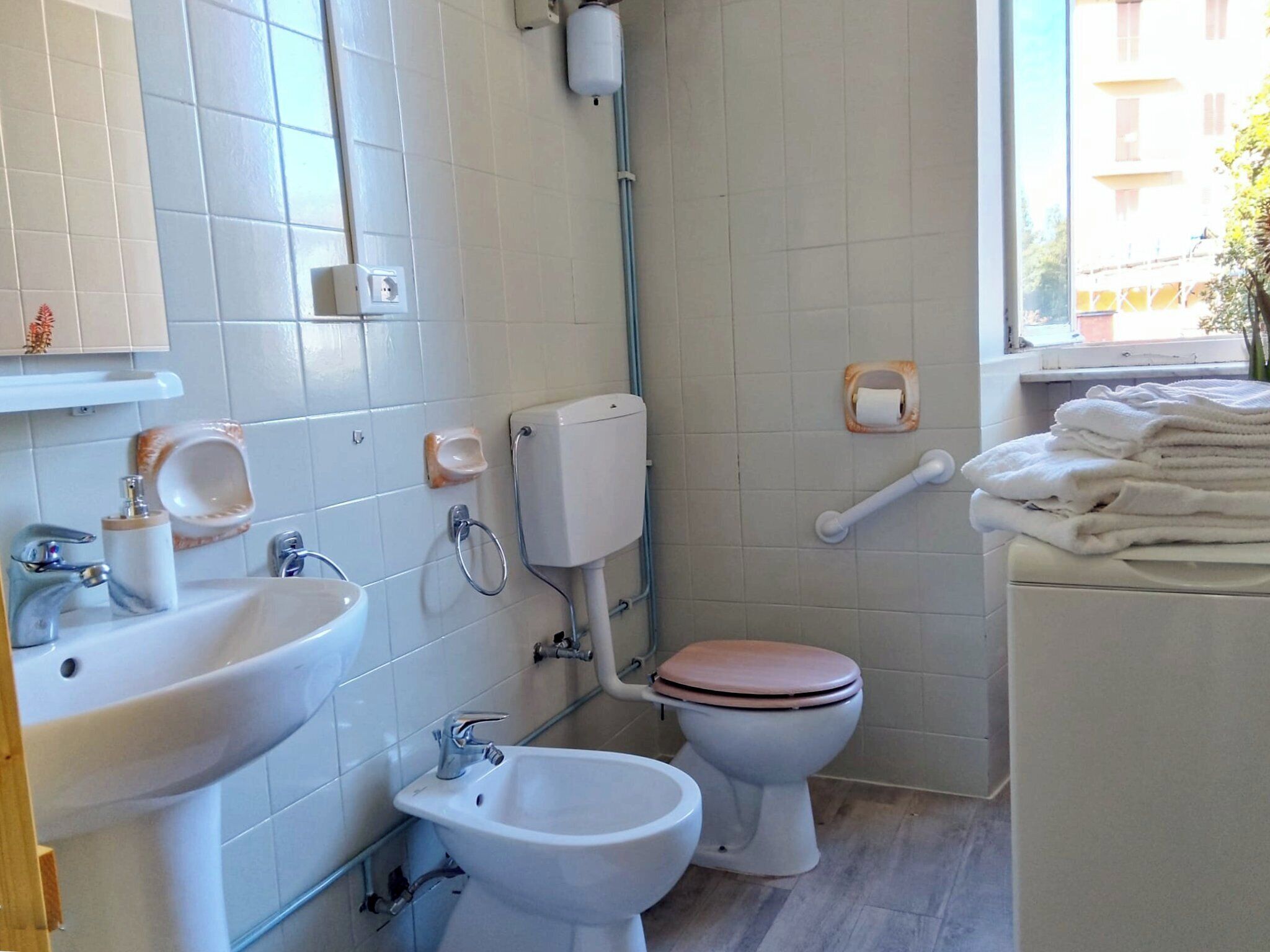 Un baño con lavamanos, bidé, inodoro y ventana con vista a edificios.