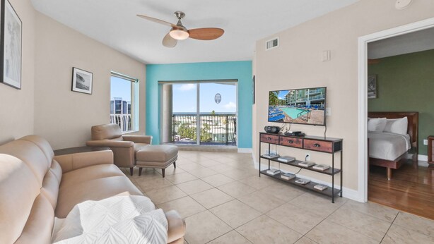 A spacious living room with a beige sofa, a coffee table, a TV on a wooden stand, and a balcony with a view of buildings and a pool. - Upscale Comfort with and Affordable Price 🏝 Beach Access across the street (Treasure Island)